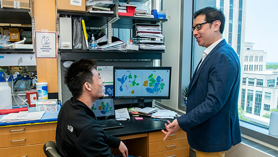 Man with glasses speaking to another man seated at a computer in a lab, with images displayed on the screen and a high-rise building in the background