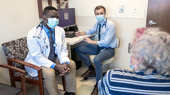 Two medical students masks, wearing white coats speaking to a woman in a mask