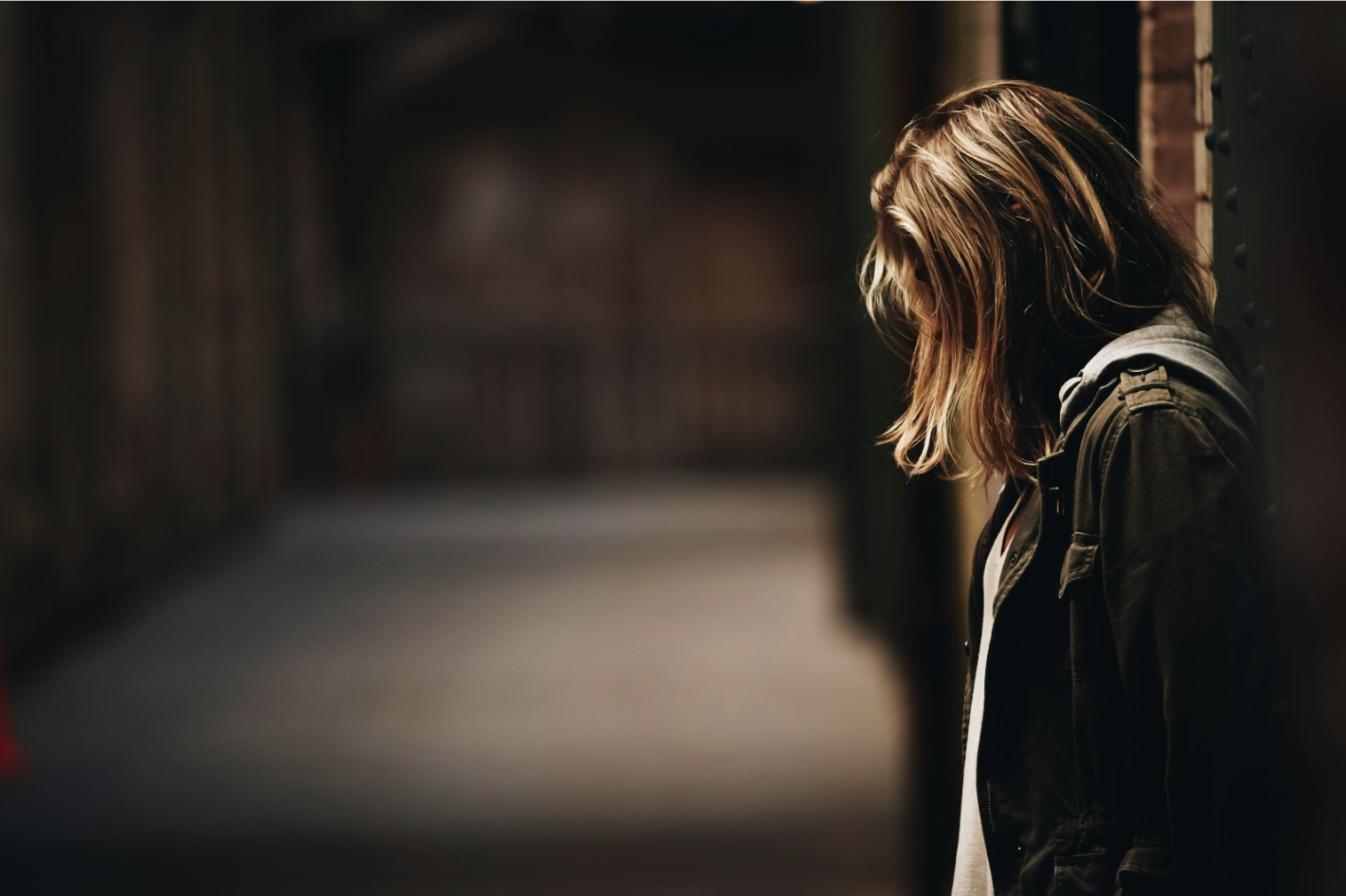 Female youth with back against wall, head down in poorly lit room. 